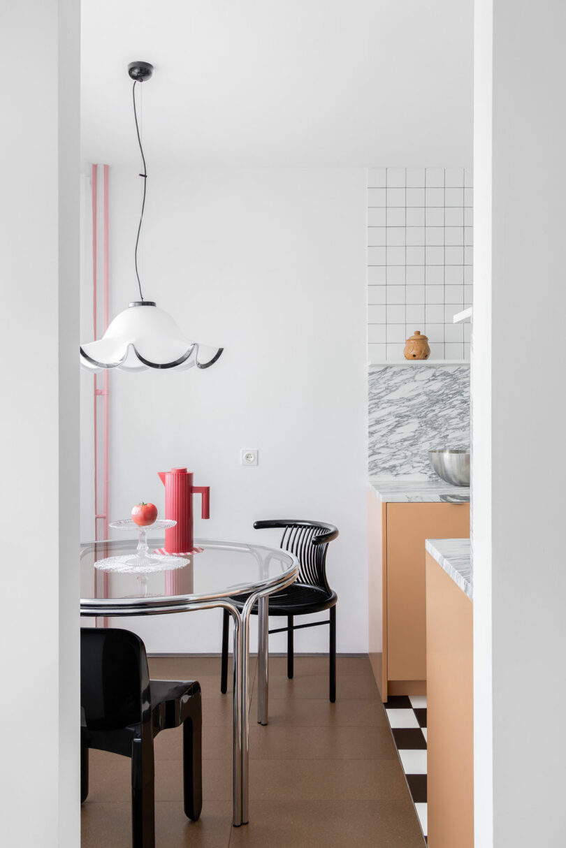 A modern kitchen with a marble countertop, peach cabinets, checkered floor, glass table, black chairs, and a white pendant light; red accents add color to the space.