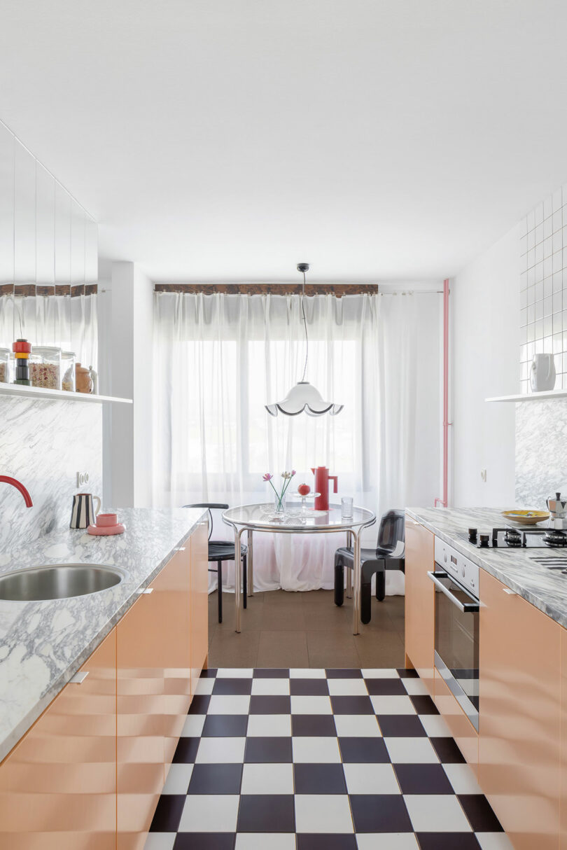Modern kitchen with peach cabinets, marble countertops, checkered floor, and a small dining area by a large window with sheer curtains. A pendant light hangs above the glass table.