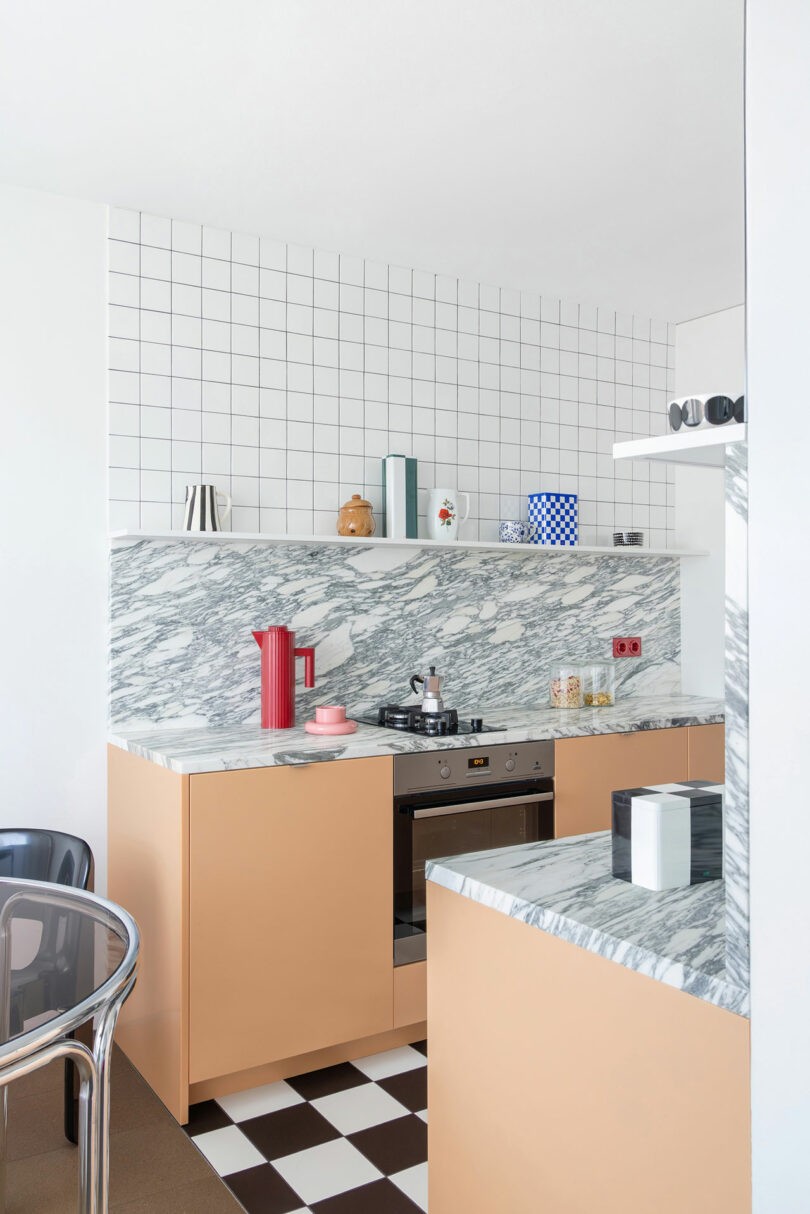 Modern kitchen with peach cabinets, marble countertops, white tile backsplash, open shelf with dishes, stainless steel oven, and checkered black-and-white floor.