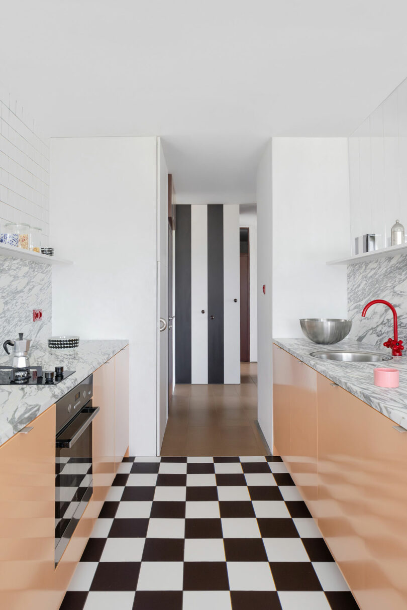 Modern kitchen with marble countertops, peach-colored cabinets, a red faucet, and a black-and-white checkered floor; a hallway with vertical stripes is visible in the background.