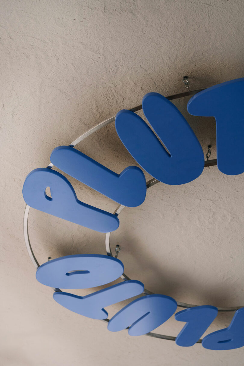 A partial view of a circular metal frame with large blue letters, possibly spelling "Pluto," mounted on it, attached to a textured beige wall.