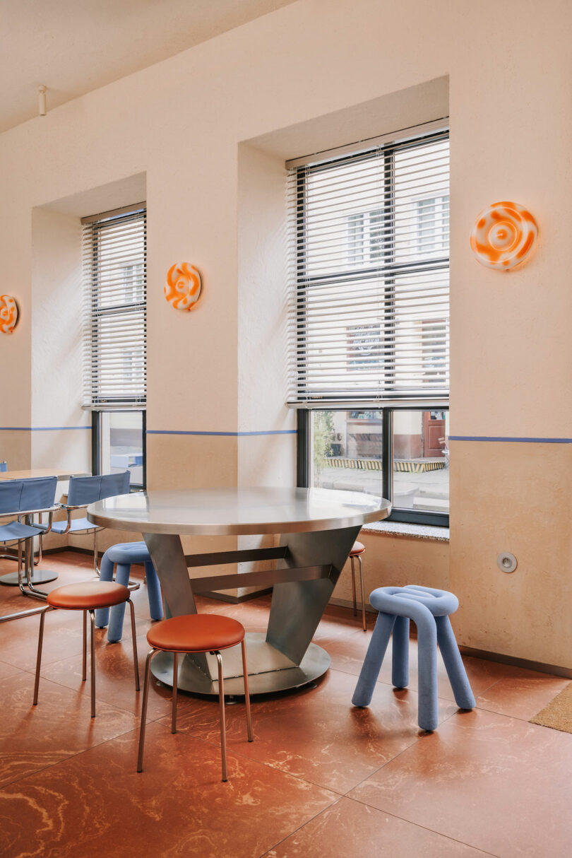 Modern café interior with a metal table, orange and blue stools, large windows with blinds, and Pluto-inspired orange wall lights on a cream-colored wall.