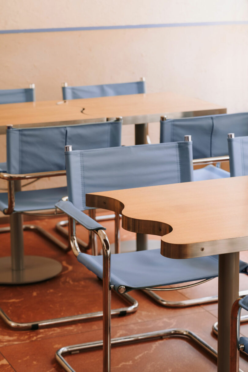 Several light wooden tables and blue fabric chairs with metal frames are arranged in a classroom or meeting room, evoking a calm atmosphere fit for learning—almost like studying the mysteries of Pluto. One table features a curved cutout on its edge.