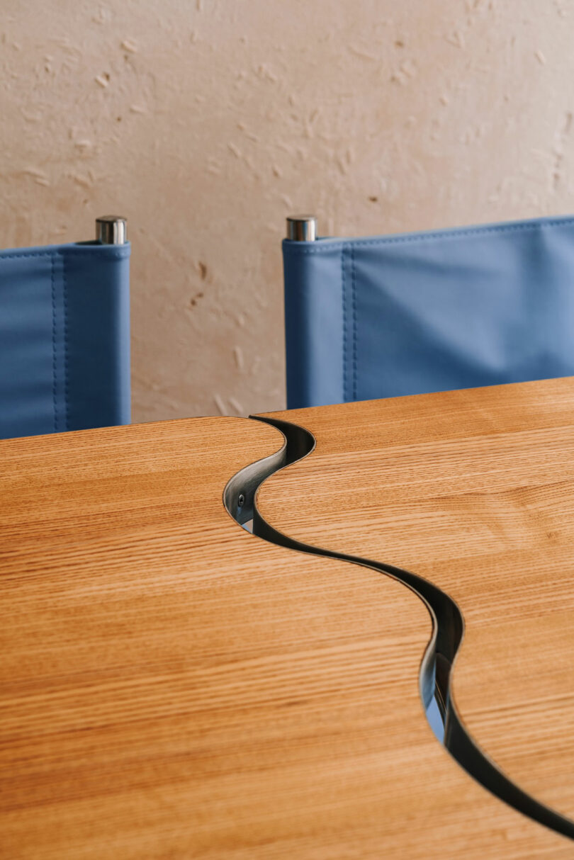 A wooden table with a wavy metal inlay reminiscent of Pluto’s icy surface runs through the center, with two blue fabric chairs positioned behind it against a textured beige wall.
