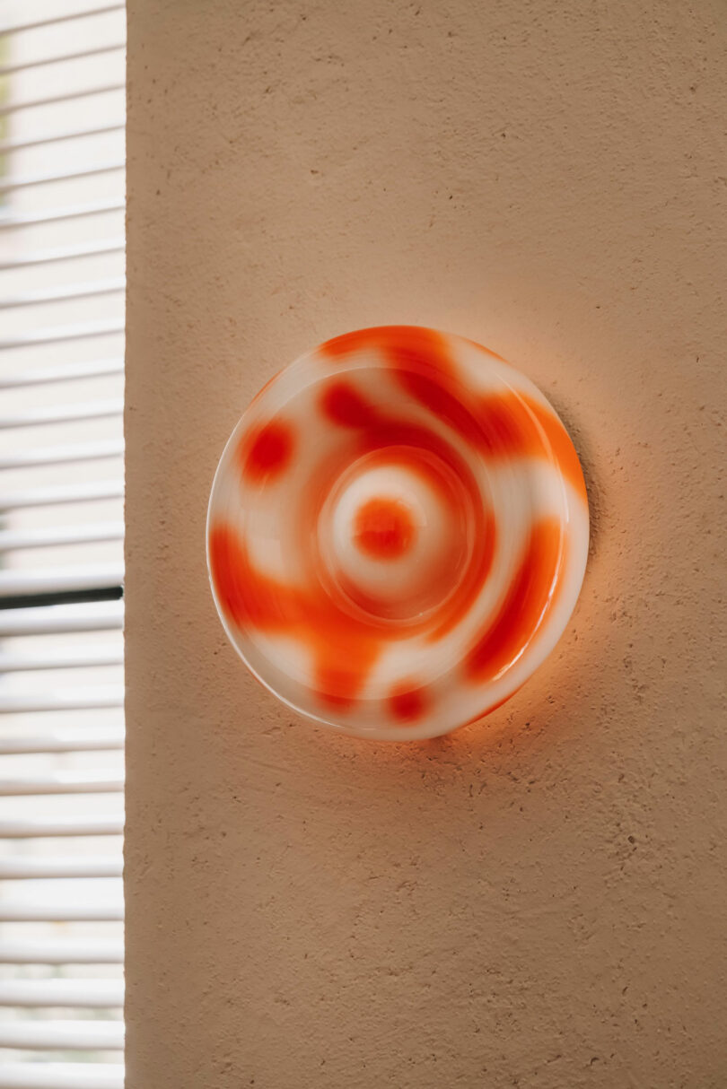 Circular Pluto-inspired wall light fixture with an orange and white swirled pattern mounted on a textured beige wall next to window blinds.