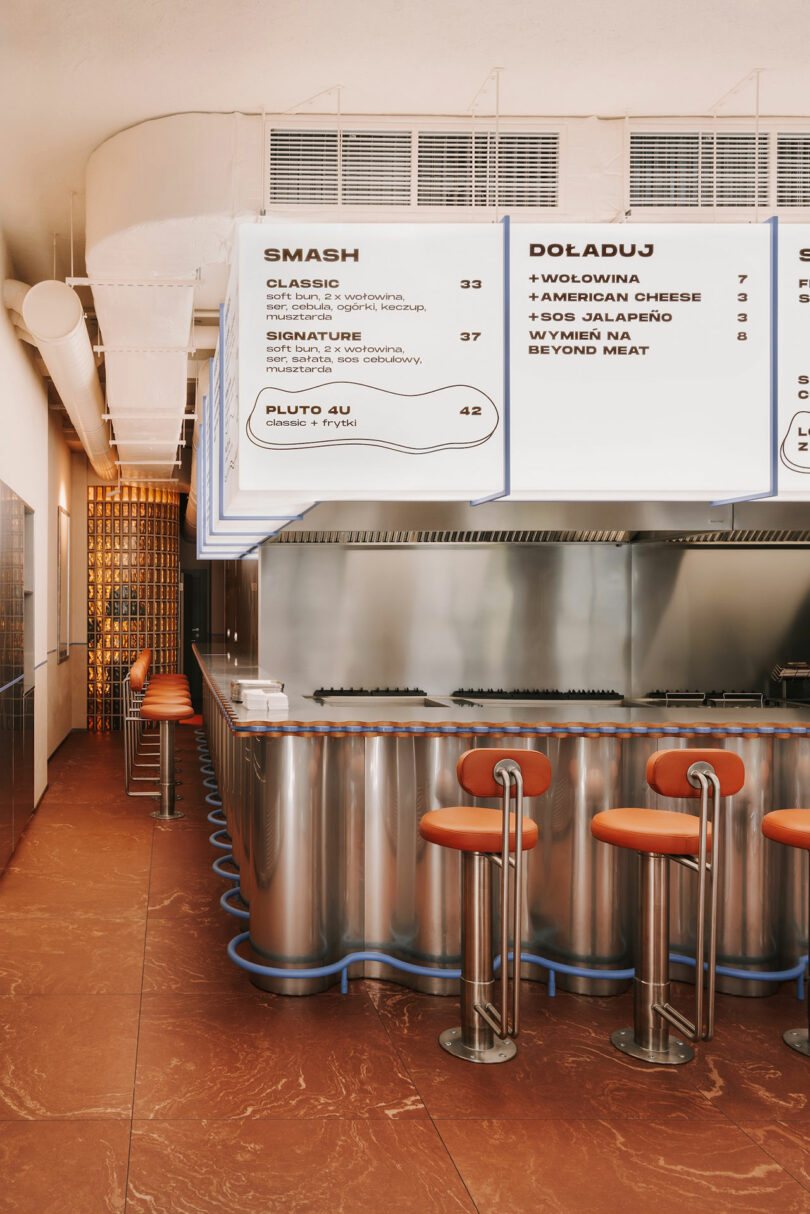 Modern diner interior with metal countertops, orange bar stools, and large Polish menu boards above the counter—featuring playful Pluto-themed accents for a unique touch.