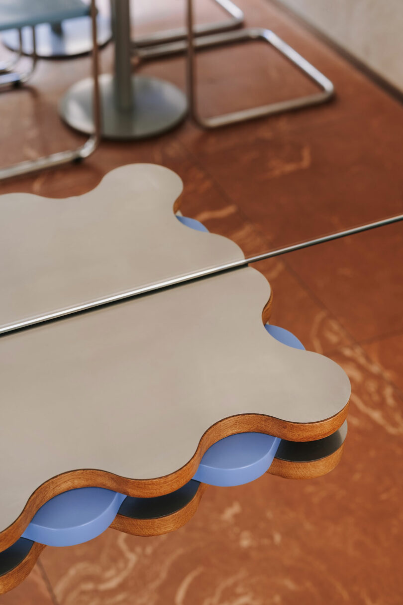 A close-up of a scalloped-edge table with a layered design, featuring wood and blue Pluto-inspired accents, seen next to metal chairs on a reddish-brown tiled floor.