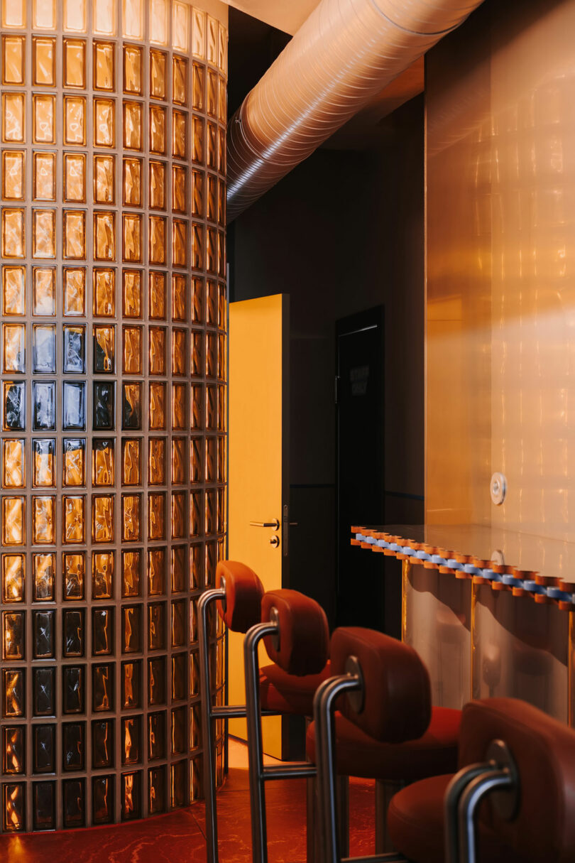 A modern interior with red bar stools, a counter, an open door, and a curved wall made of glass blocks illuminated by warm lighting creates an atmosphere that's as inviting as Pluto’s distant allure.