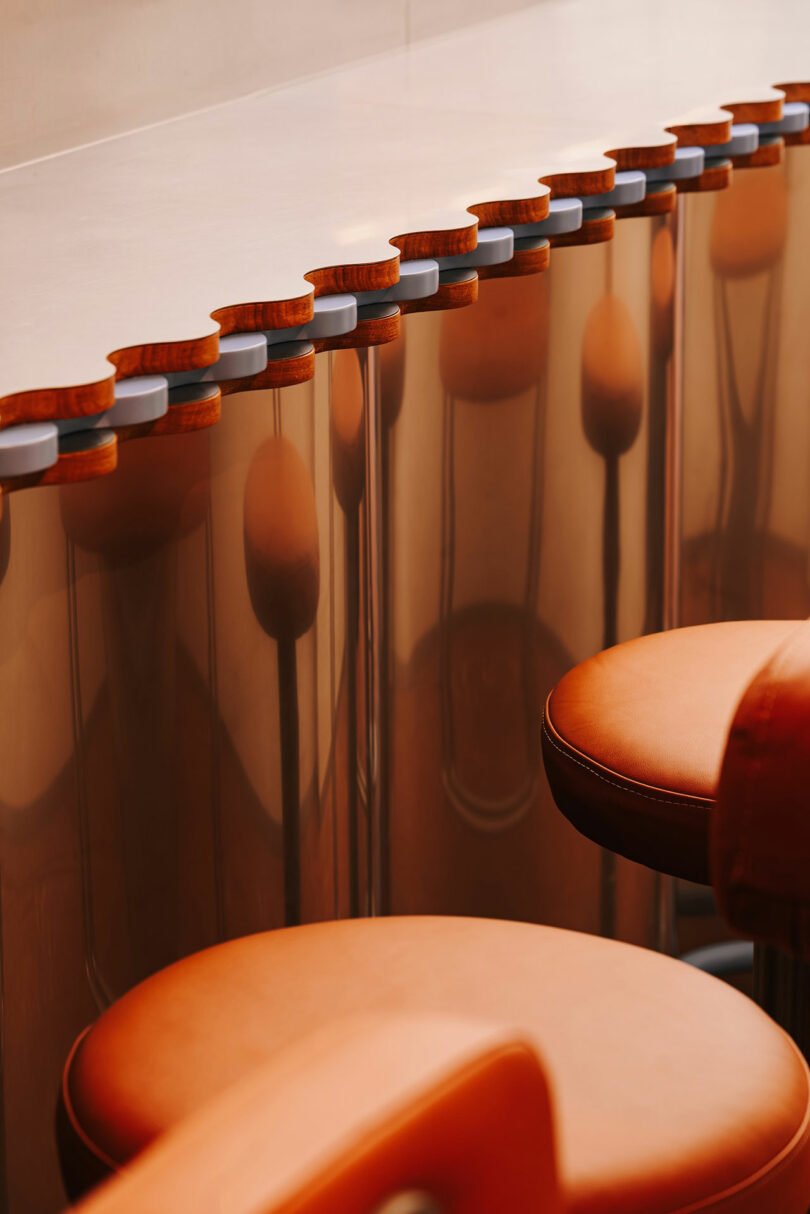 A row of orange leather bar stools with rounded backs, reflecting on a polished metallic counter beneath a smooth surface, creates a sleek vibe fit for Pluto’s coolest lounge.