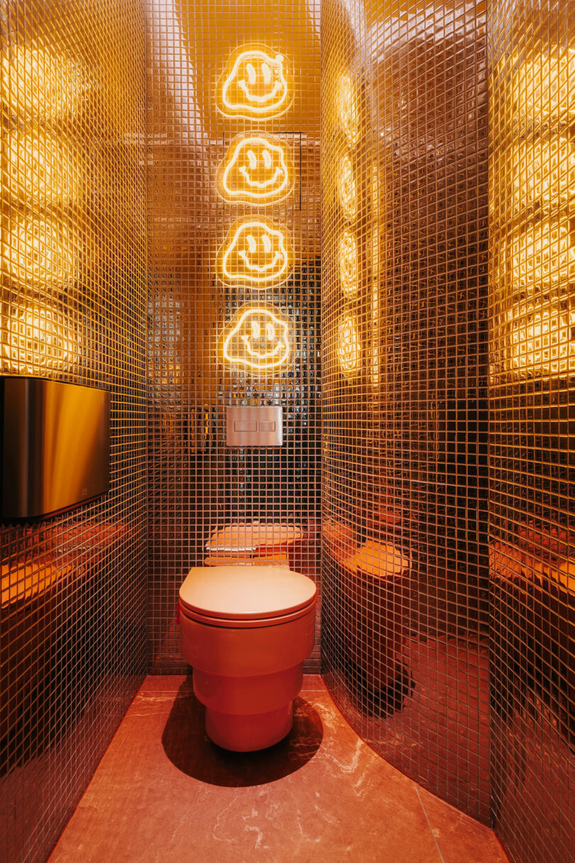 A modern bathroom with metallic mosaic tile walls, a wall-mounted toilet, and yellow neon cloud lights above the toilet creates a celestial vibe that feels straight from Pluto.
