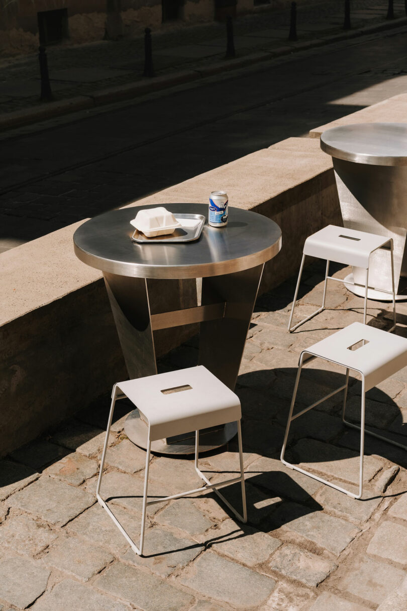 A round metal table with two white stools on a stone-paved sidewalk, holding a tray, takeout container, and a can of Pluto drink.