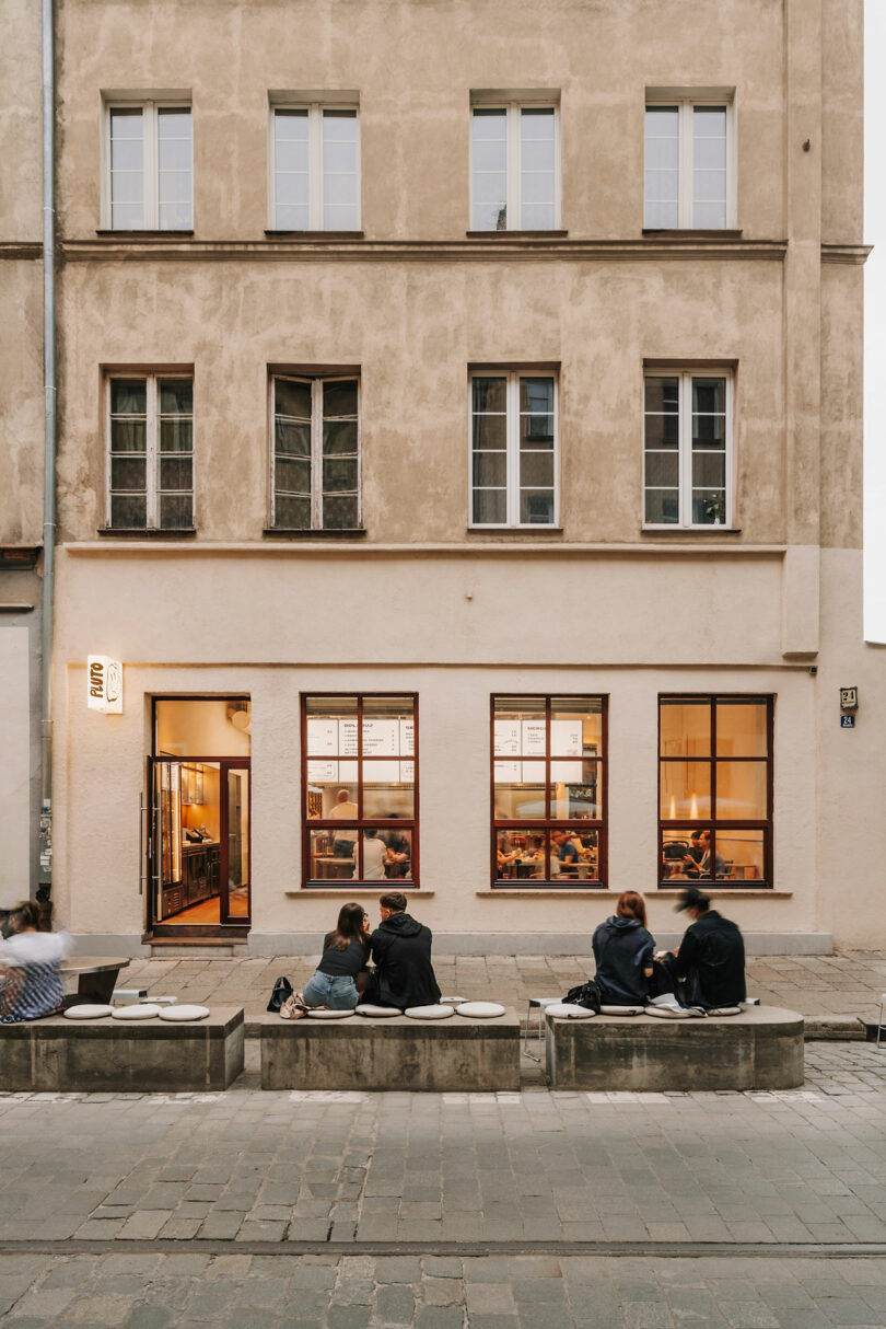 A beige, three-story building with large windows and an open door on the ground floor sits beneath a clear sky—several people lounge on concrete benches outside, chatting as if Pluto itself were just another neighbor.