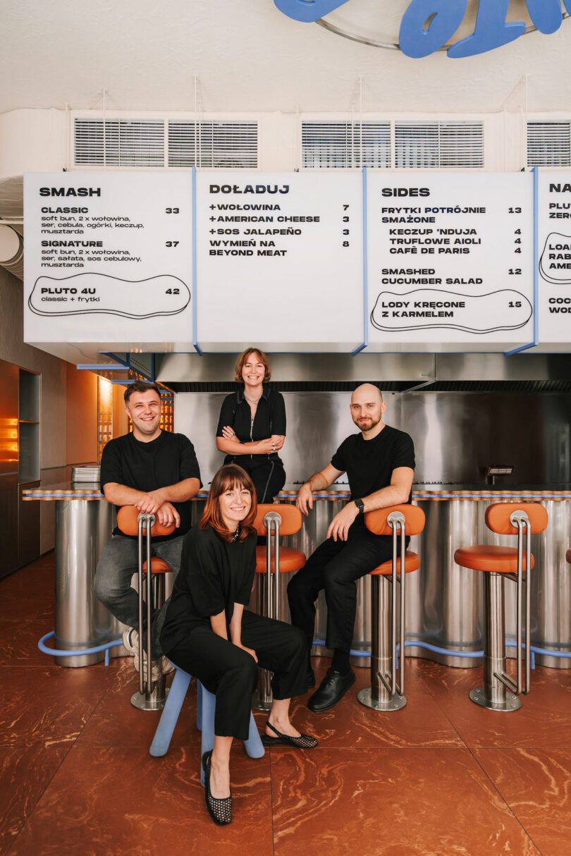 Four people dressed in black pose and smile inside Pluto, a modern restaurant, under a large menu board displaying food and drink options.