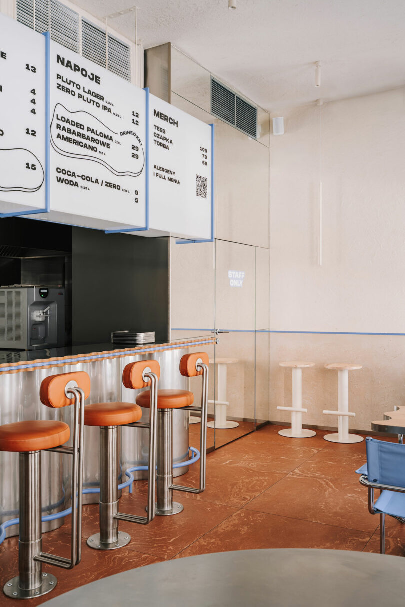 Modern cafe interior with orange bar stools at a counter, a Pluto-themed menu board above, blue seating against the wall, and a staff-only door in the background.