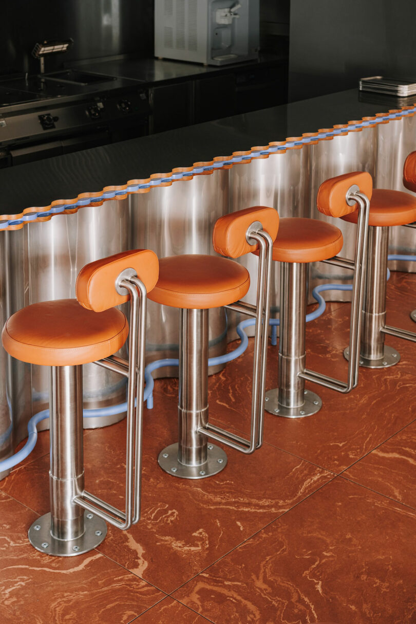 Four orange bar stools with metal bases are lined up at a shiny metallic counter with blue Pluto-blue accents, on a reddish-brown tiled floor in a modern setting.
