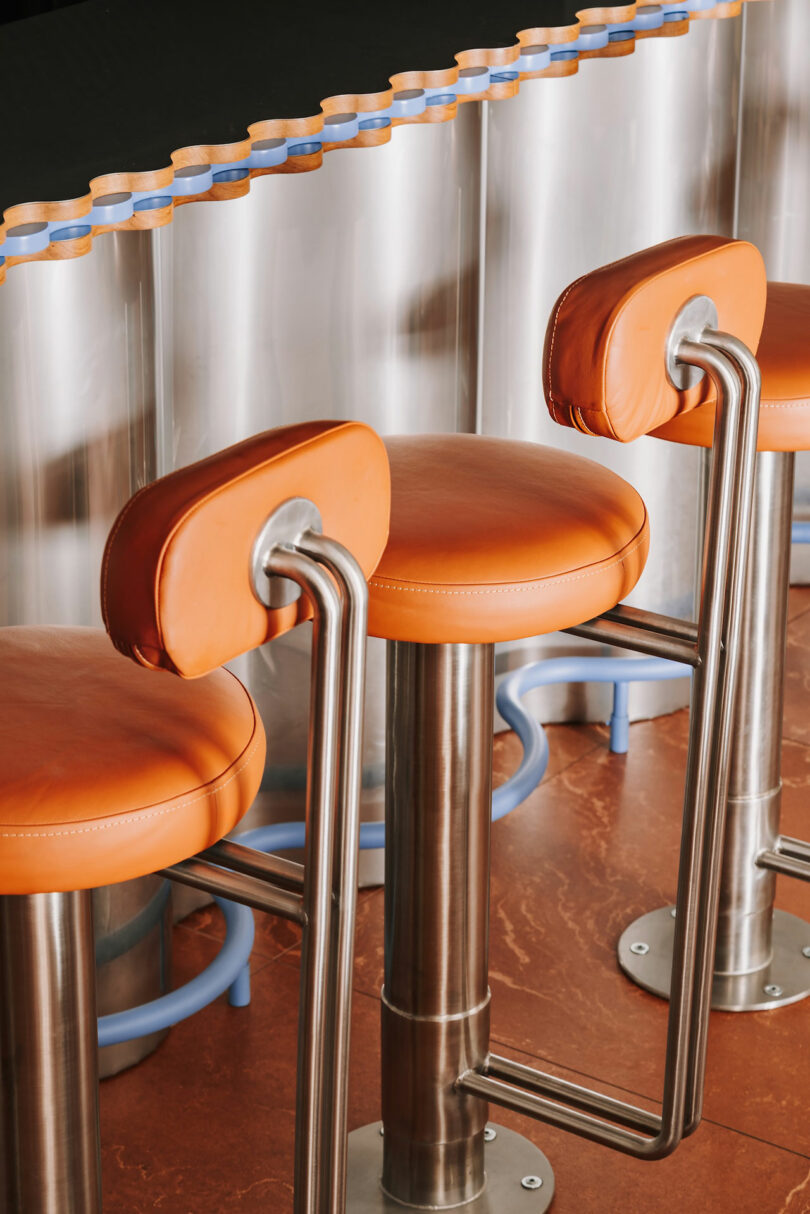 Three orange leather barstools with metal frames are lined up in front of a counter with a silver metallic and wavy Pluto-inspired design.