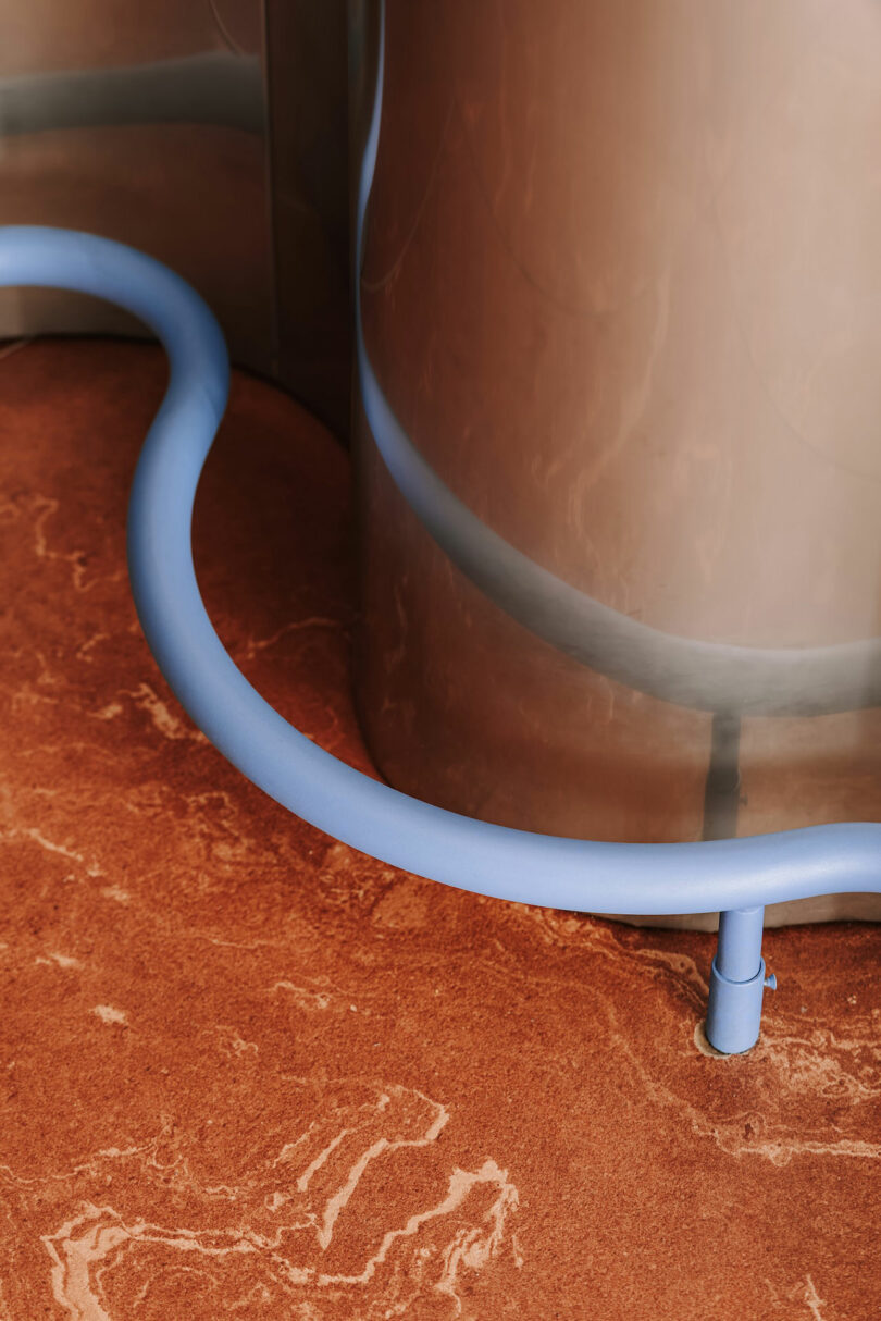 A blue hose is connected to a large, metallic cylindrical tank, resting on a reddish-brown, marbled floor that evokes the distant surface of Pluto.