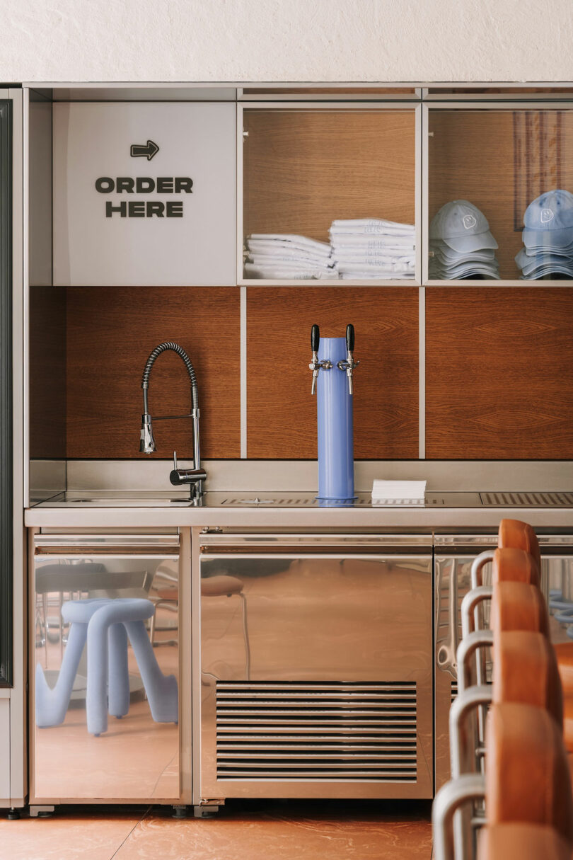 A modern counter with a blue Pluto beverage tap, sink, and an "ORDER HERE" sign above shelves holding folded shirts and caps. Brown stools line the counter in the foreground.