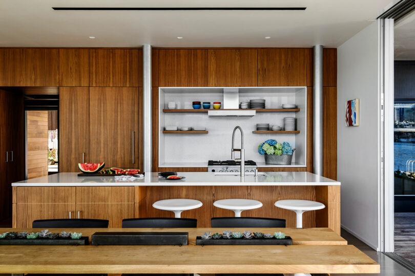 Cocina moderna con gabinetes de madera, encimeras blancas, estanterías abiertas y taburetes; Una mesa de comedor con suculentas está en primer plano.
