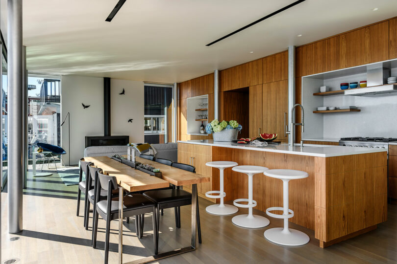 Moderna cocina y comedor de planta abierta con gabinetes de madera, una gran isla con taburetes de bar blanco, una mesa de comedor de madera y sala de estar en el fondo.