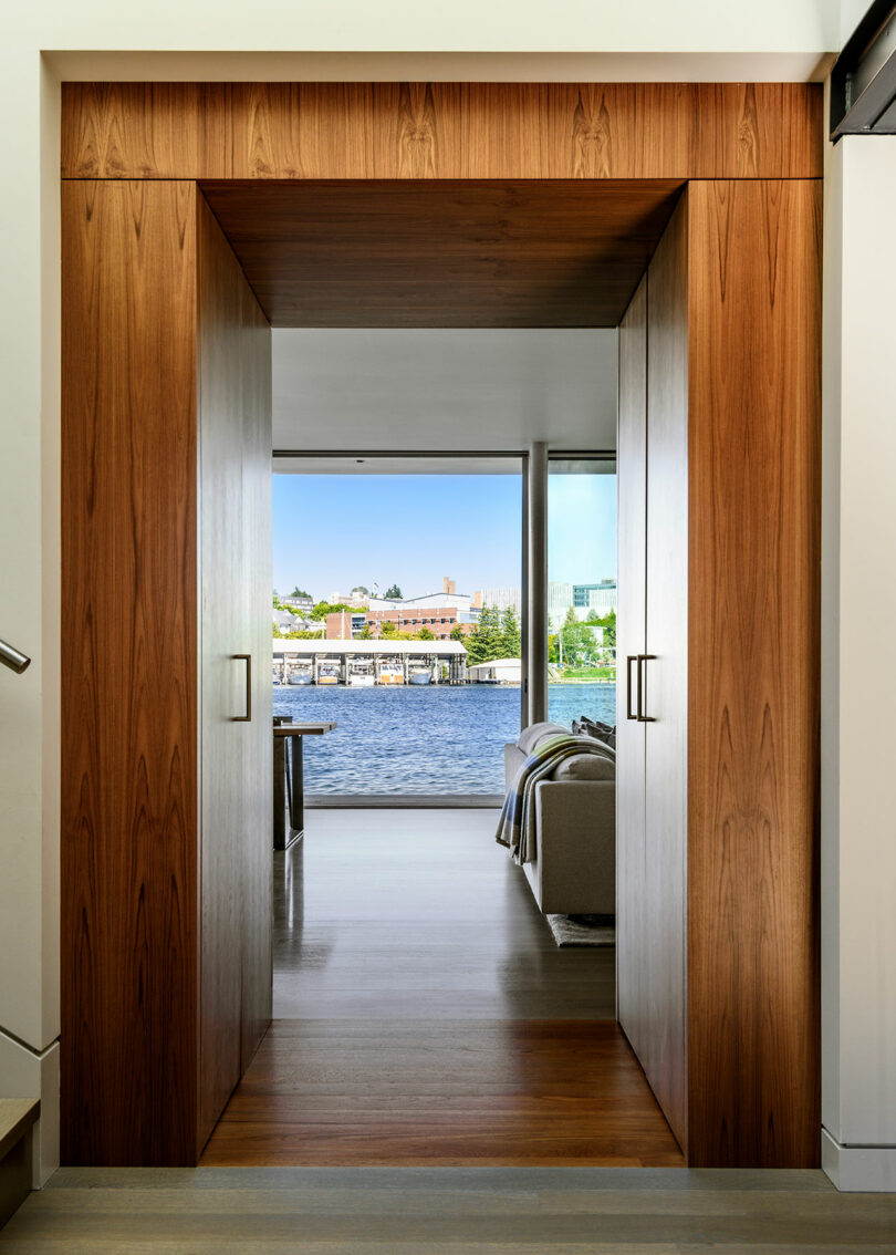 Una puerta de madera enmarca una vista de una sala de estar moderna con un sofá y una gran ventana con vistas al agua y los edificios afuera.