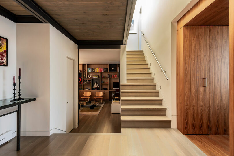 Interior moderno con una escalera de madera, paredes blancas, almacenamiento de madera incorporado y una sala de estudio adyacente con estanterías y muebles de estilo de mediados de siglo.