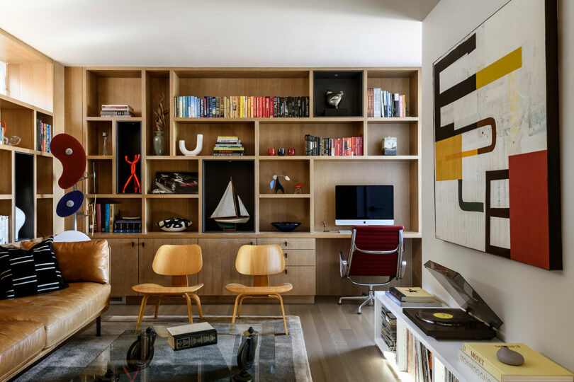 Moderna oficina en casa con estanterías de madera, libros coloridos, un iMac en un escritorio, dos sillas de madera, arte abstracto, un tocadiscos y un sofá de cuero marrón.