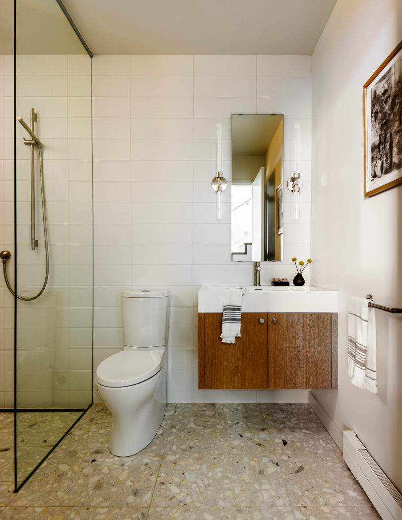 Baño moderno con un inodoro blanco, tocador de madera, espejo rectangular, recinto de ducha de vidrio y paredes de azulejos blancos. Una toalla cuelga en el fregadero y el arte está montado en la pared.