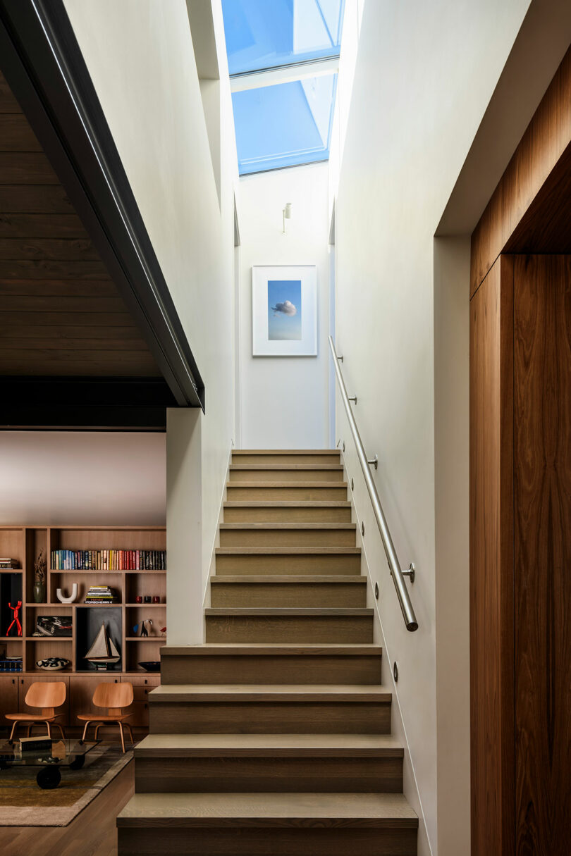 Una moderna escalera de madera con un pasamanos de metal conduce a una claraboya; Una imagen enmarcada cuelga en la pared, y una sala de estar con estantes es visible a la izquierda.