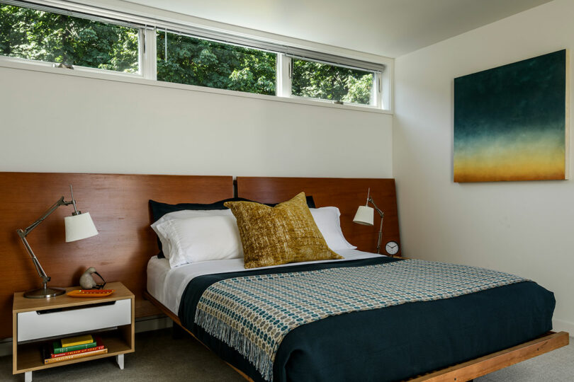 Un dormitorio moderno con cama de madera, ropa de cama azul, una almohada de acento amarillo, dos mesas de noche con lámparas y una gran pintura abstracta en la pared.