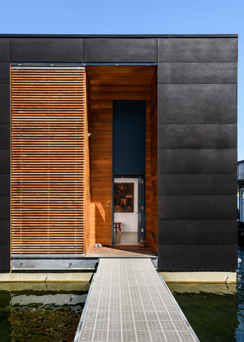 Moderna casa flotante con paneles exteriores oscuros y listones de madera, vistas desde una pasarela que conduce a una entrada abierta sobre el agua.