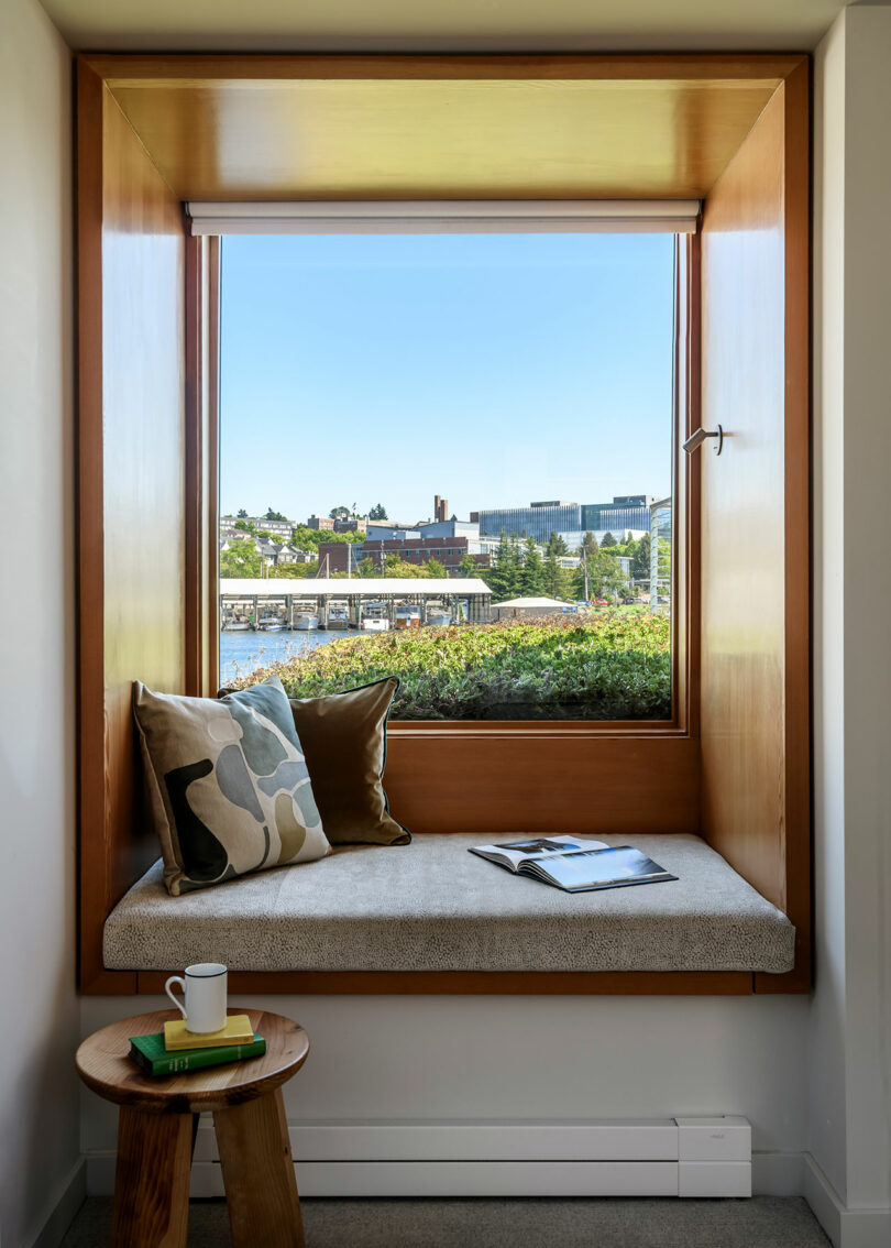 Un asiento de ventana incorporado con dos almohadas y un libro abierto da a una vista frente al mar; Una pequeña mesa lateral contiene una taza y dos libros.