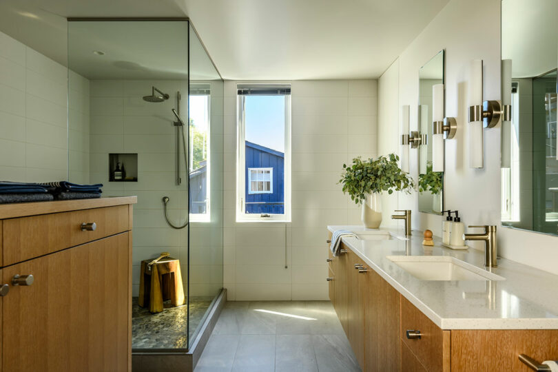 Baño moderno con ducha de vidrio, gabinetes de madera, tocador de doble fregadero, espejo grande, planta en maceta y una ventana que deja en luz natural.