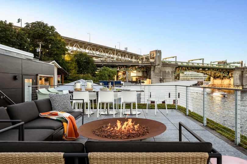 Moderno patio al aire libre con un pozo de fuego, sofá negro, asientos de bar y un comedor con vistas a un río y un puente cercano bajo cielos despejados.