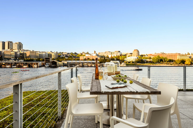 Mesa de comedor al aire libre con sillas blancas, cristalería y limas en una terraza con vistas a un río tranquilo y un paisaje urbano debajo de un cielo azul claro.