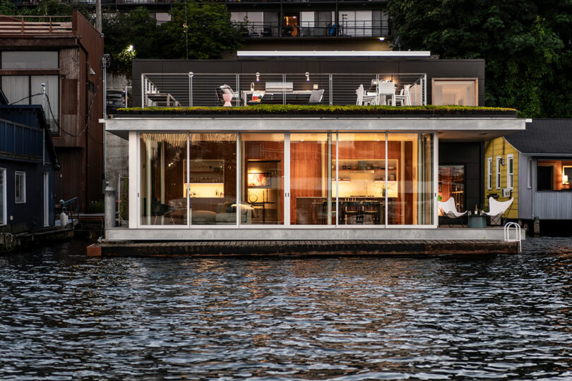 Moderna casa flotante con grandes ventanas de vidrio, área de asientos en la azotea y luces interiores encendidas, situada en un cuerpo de agua con árboles y edificios en el fondo.