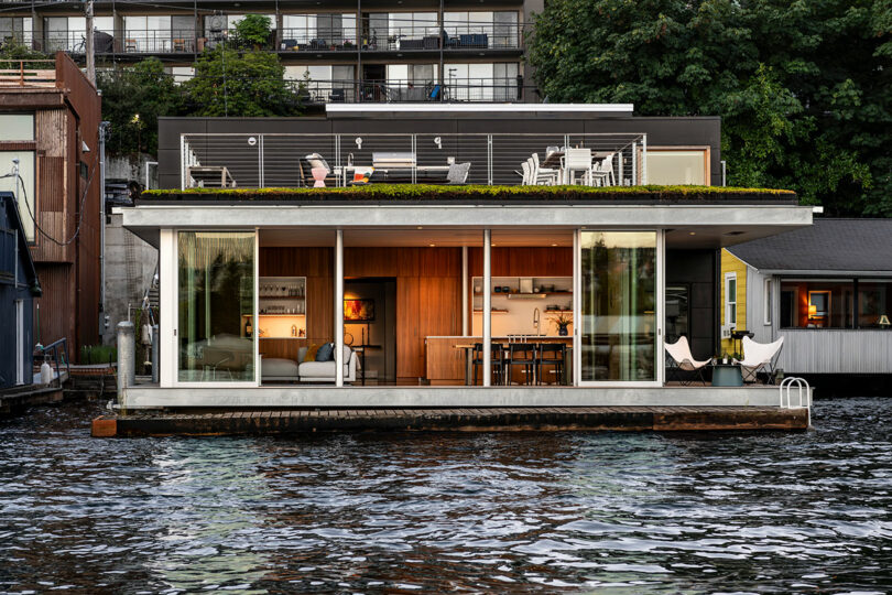 Una casa flotante moderna con grandes ventanas de vidrio y una cubierta de la azotea se acopla en un cuerpo de agua, con edificios vecinos y vegetación en el fondo.