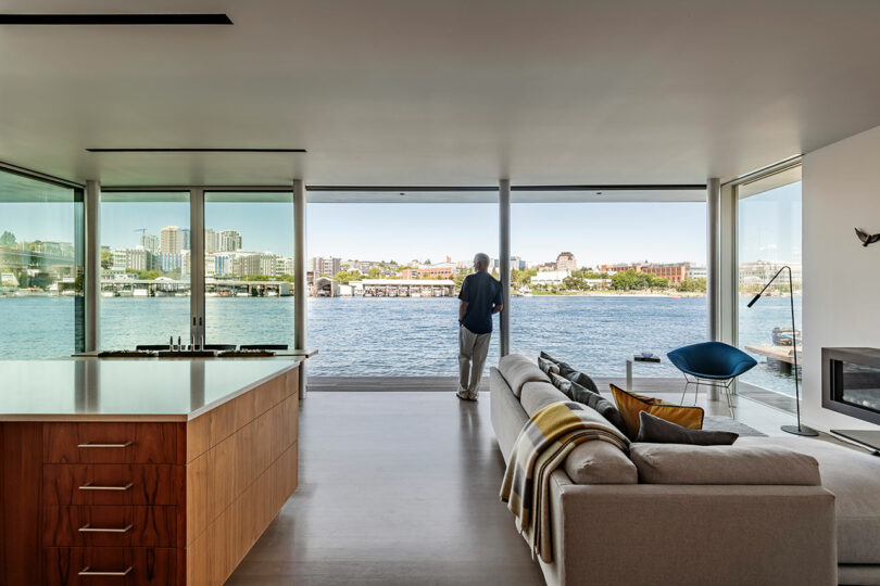 Una persona se encuentra con grandes ventanas en una sala de estar moderna, con vistas a un paisaje urbano frente al mar en un día despejado.