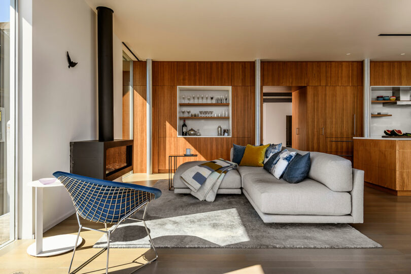 Sala de estar moderna con paredes con paneles de madera, un sofá seccional gris con almohadas coloridas, una alfombra gris, una estufa de madera negra y una silla de acento azul cerca de grandes ventanas.