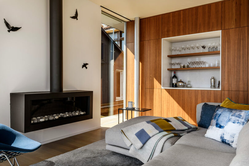 Sala de estar moderna con chimenea negra, decoración de la pared de pájaros, área de barra de madera incorporada con cristalería y un sofá seccional gris con manta y almohadas.