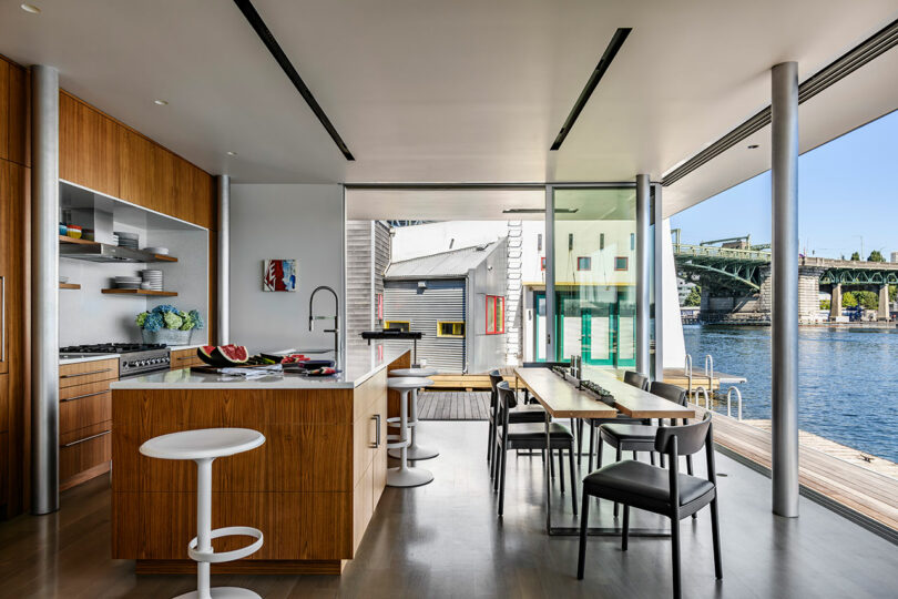 Cocina moderna y comedor con acabados de madera, ventanas grandes y una vista de un río y un puente afuera.