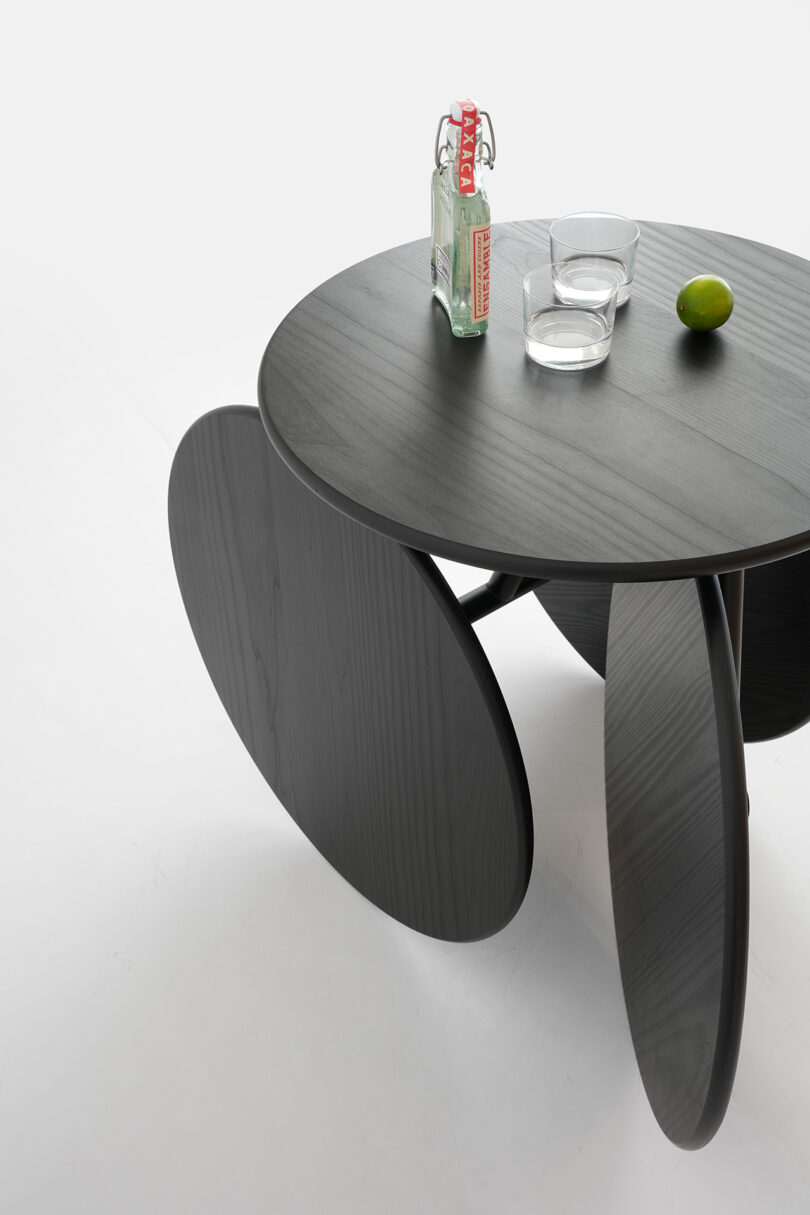 Una mesa redonda negra con patas de forma singular, sobre la que hay una botella de vidrio, dos vasos vacíos y un trozo de lima en la superficie.