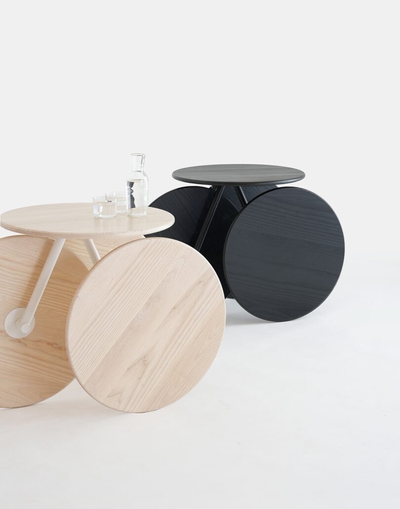 Two round tables with large circular bases, one in light wood and one in black, placed on a white background. The light wood table holds a glass carafe and two glasses.