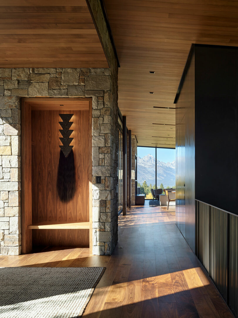 Un pasillo moderno con paredes de piedra y madera, pisos de madera, un banco con arte de pared y grandes ventanas con vistas a las montañas en el fondo.