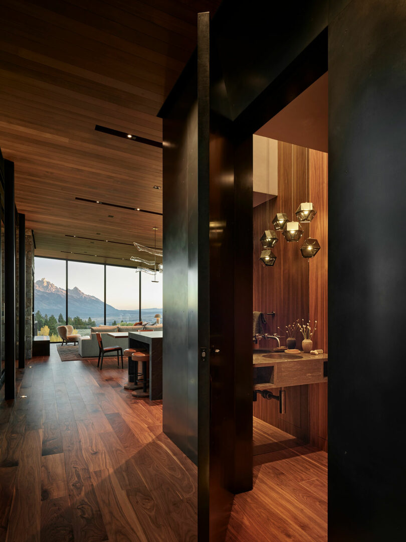 Pasillo interior moderno con pisos de madera y techo, abierto a una sala de estar con vistas a la montaña; Un baño con luces colgantes es visible a la derecha.