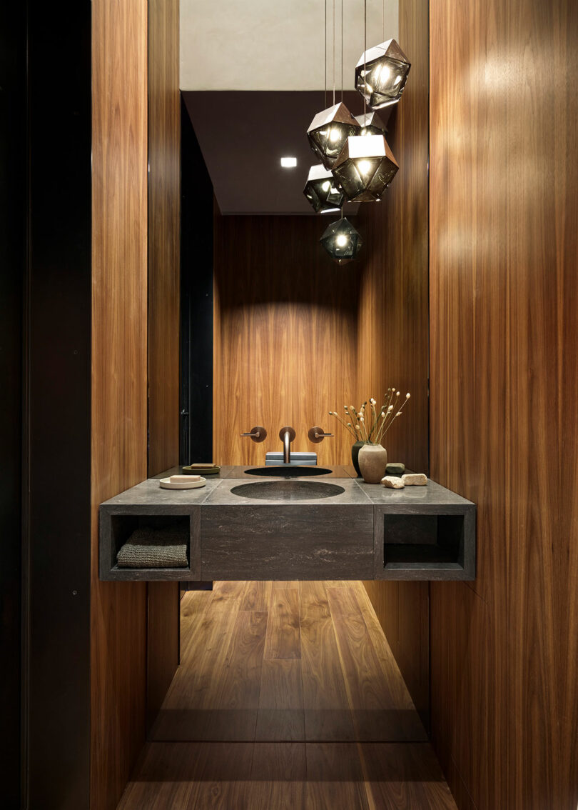 Baño moderno con paredes con paneles de madera, tocador de fregadero negro flotante, luces colgantes geométricas y decoración minimalista.