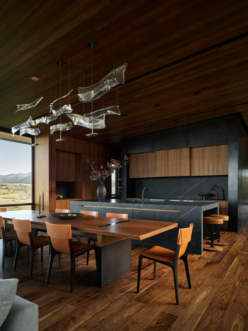 Cocina moderna y comedor con pisos de madera, paredes y techos con paneles de madera, encimeras negras, sillas de bronceado y luces colgantes abstractas, con grandes ventanas que muestran una vista al aire libre.