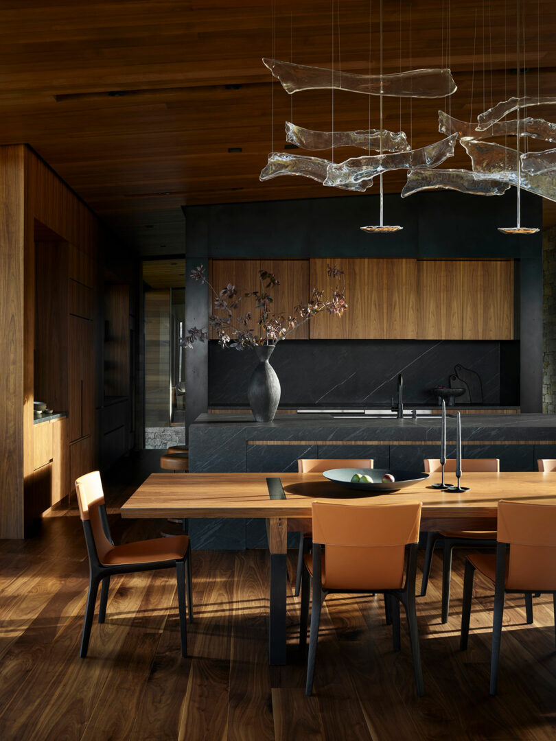Cocina moderna y comedor con pisos de madera, gabinetes y mesa, sillas de naranja, encimeras de piedra oscura y luces colgantes abstractas de vidrio arriba.