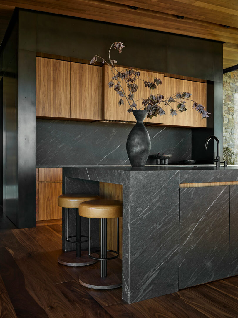 Cocina moderna con encimeras de piedra oscura e isla, gabinetes de madera, dos taburetes y un jarrón negro con ramas secas en la isla.