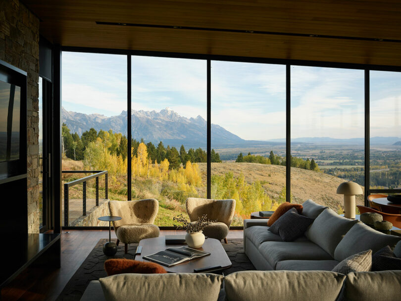 Sala de estar moderna con grandes ventanas de vidrio que ofrece una vista panorámica de montañas, árboles y un paisaje abierto. Los muebles y la decoración contemporáneos están dispuestos por dentro.